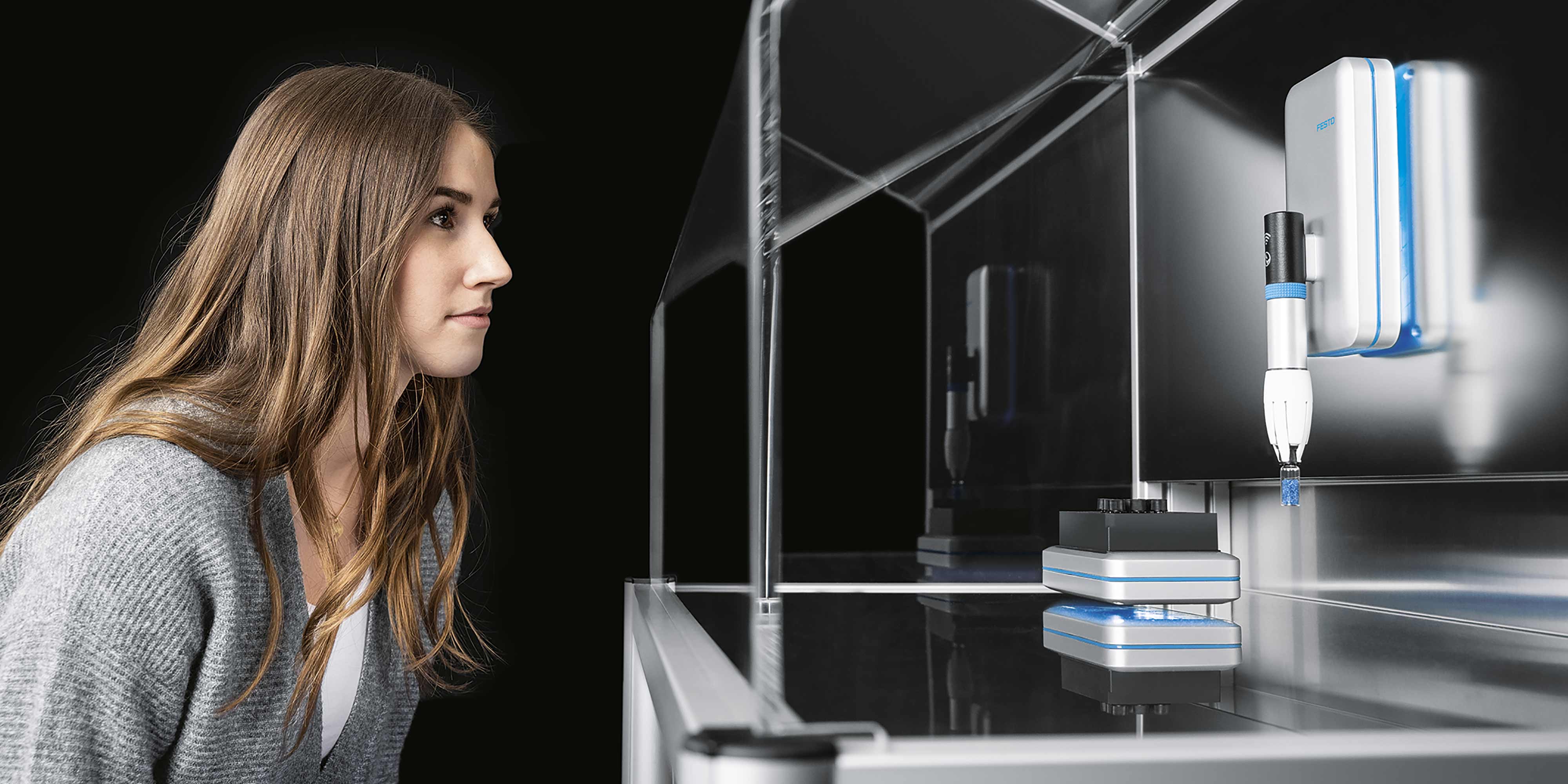 Junge Frau mit braunen Haaren schaut in einen Glaskasten mit einem weißen Greif-Modul.